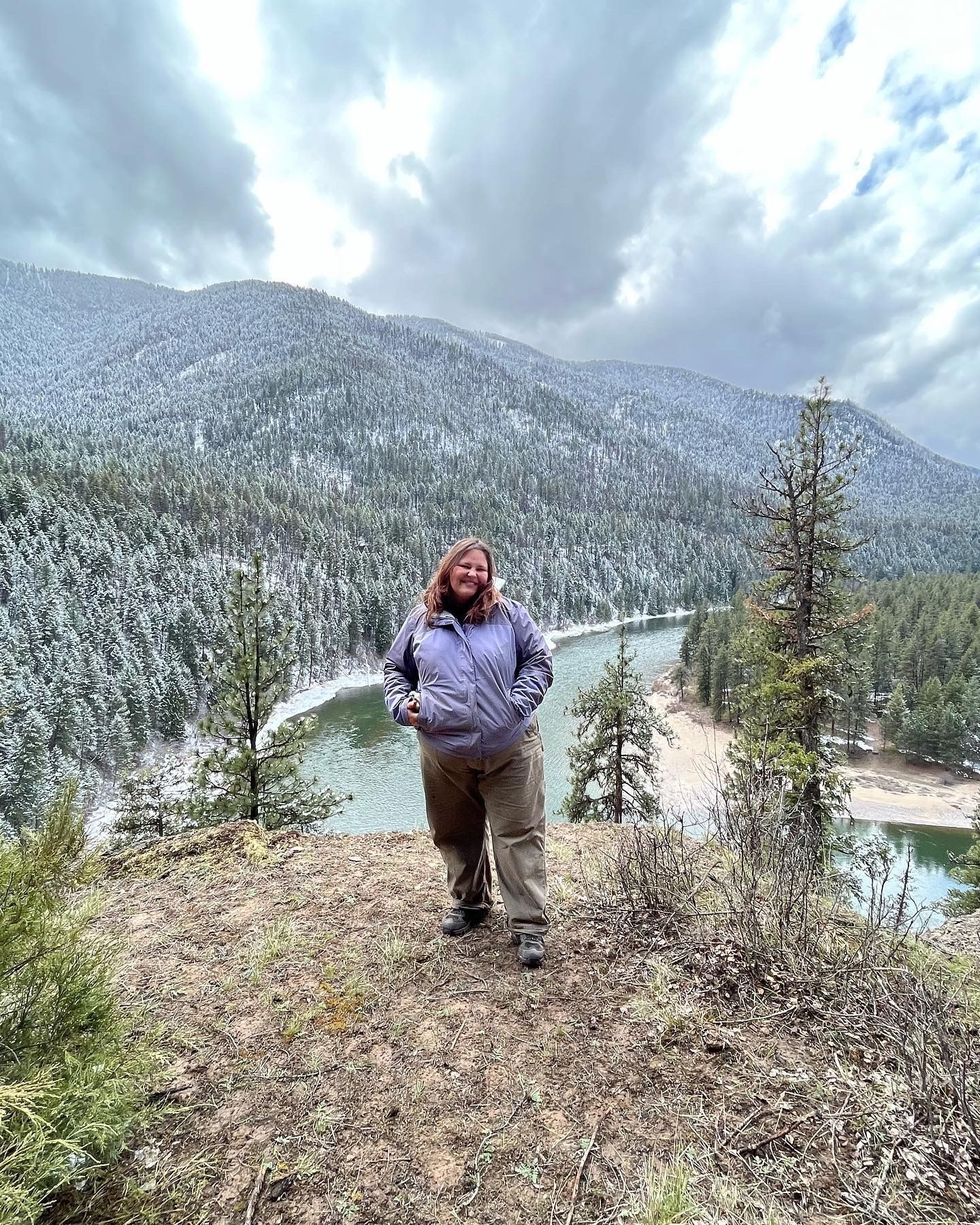 A women stands on an overlook. Below her is a river and behind her are snow dusted trees.