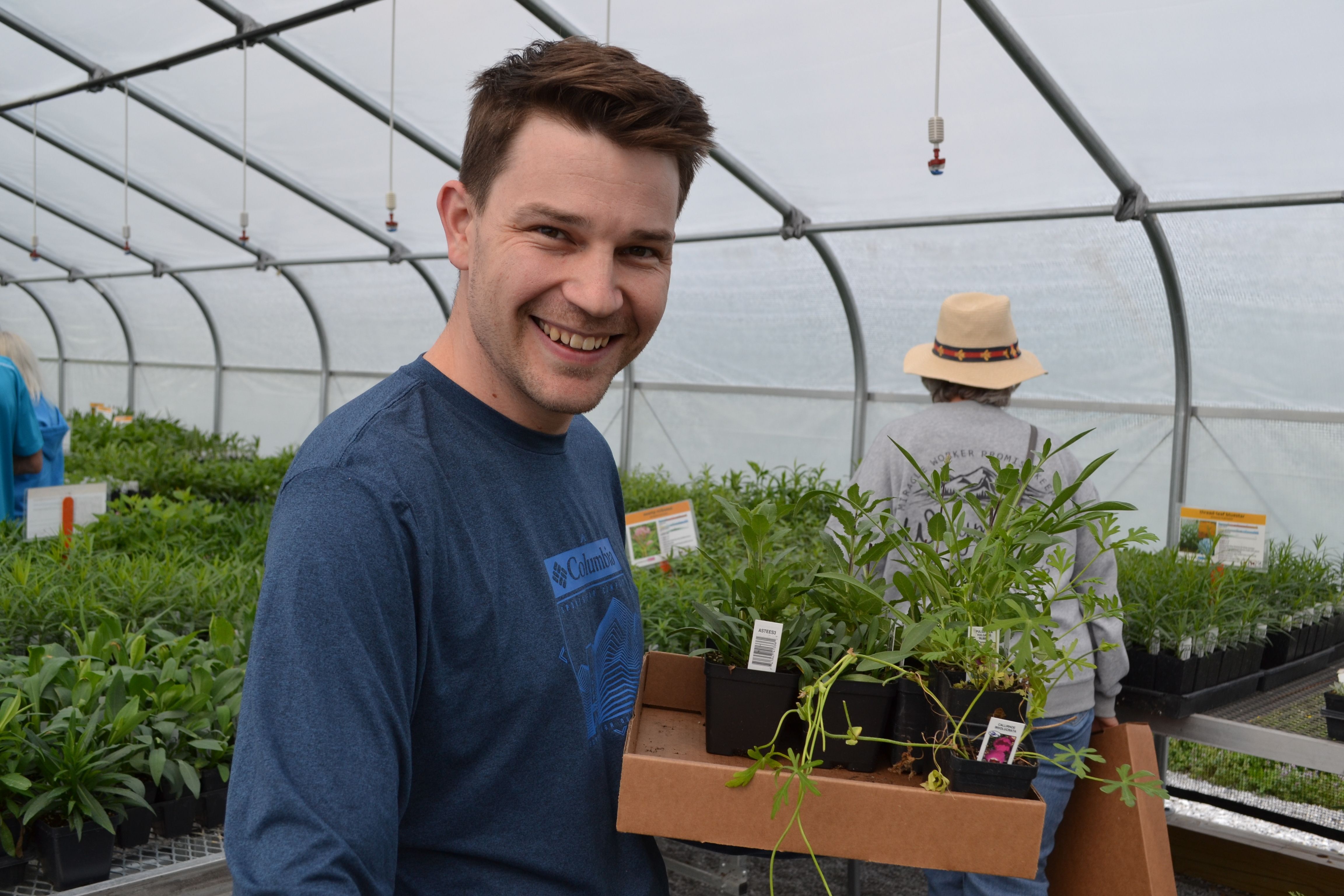 Man wearing a blue shirt holds a cardboard box of plants in a greenhouse.