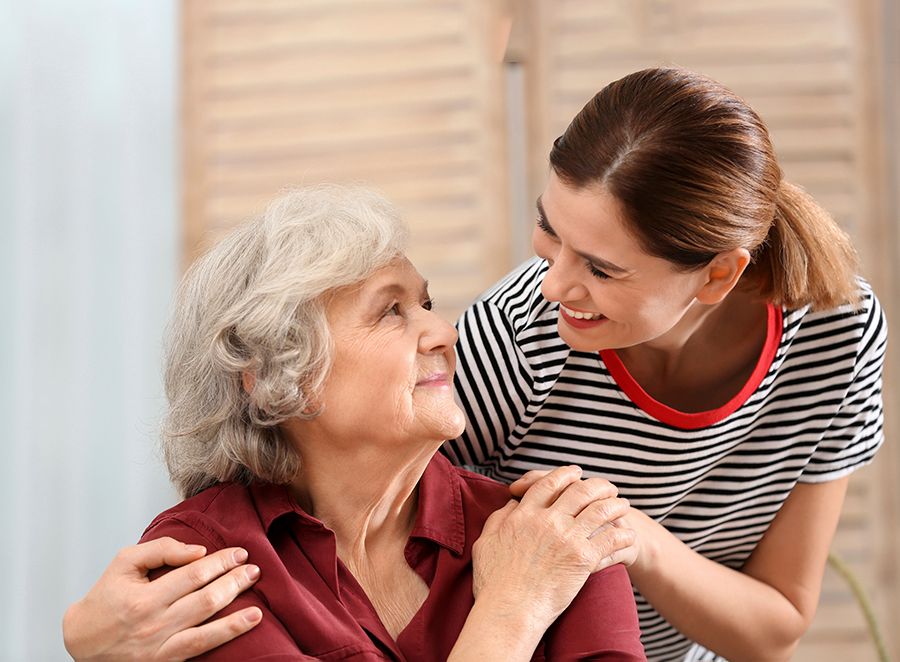 Older woman sitting at home, looking up at a younger woman who has her arm on her shoulder
