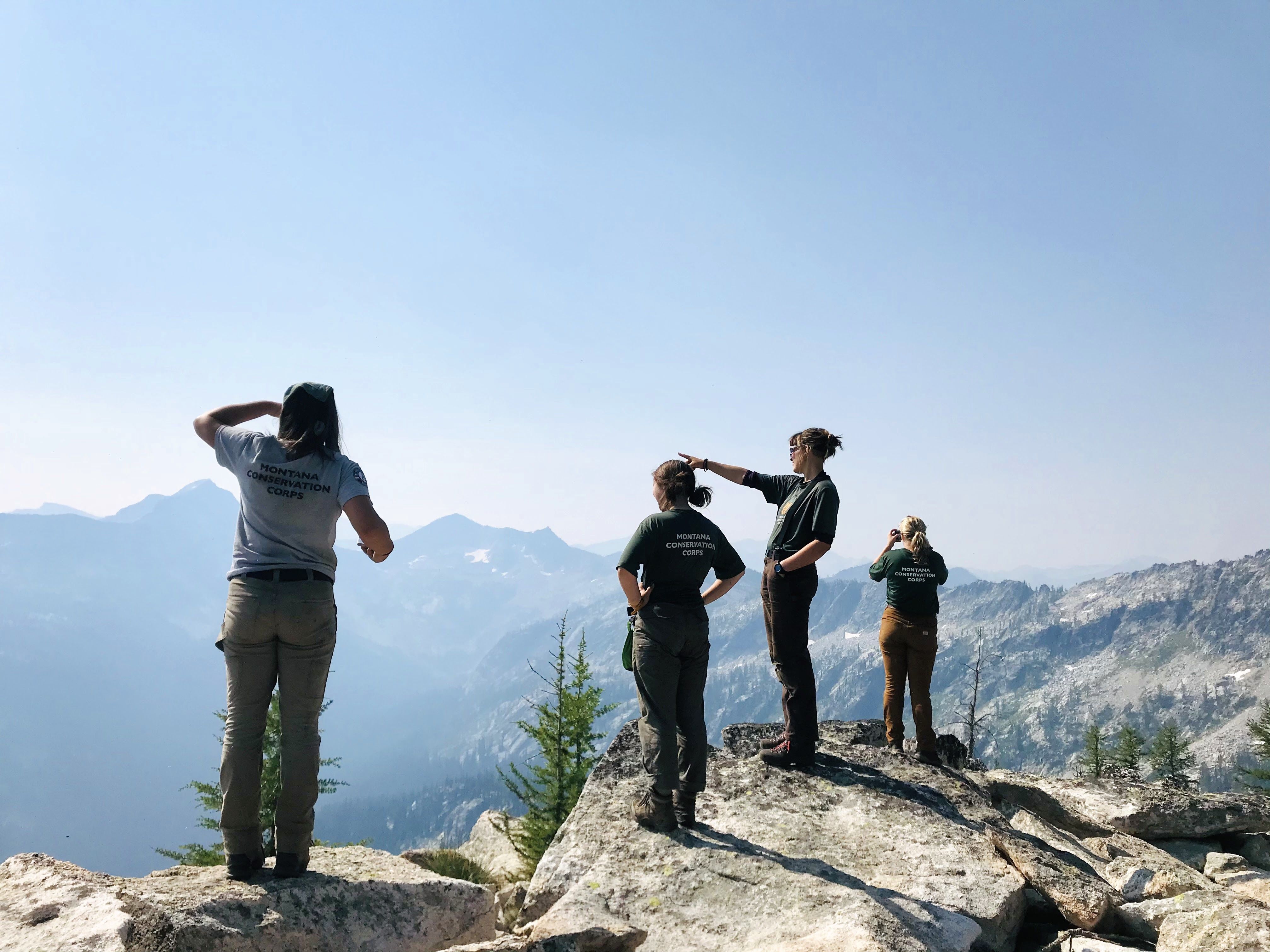 [Image Description: Four MCC members standing on a rock, looking out in the distance at the mountain ranges ahead of them. On the far right, one member is taking a photo.]