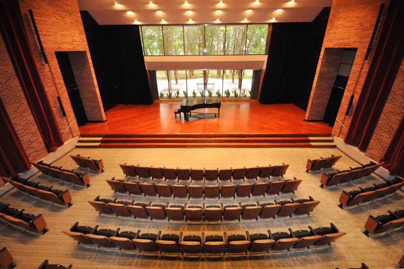 The empty Stella Boyle Smith Concert Hall at UA Little Rock. A grand Piano sits on a stage with large glass windows behind it overlooking the garden outside the building. 