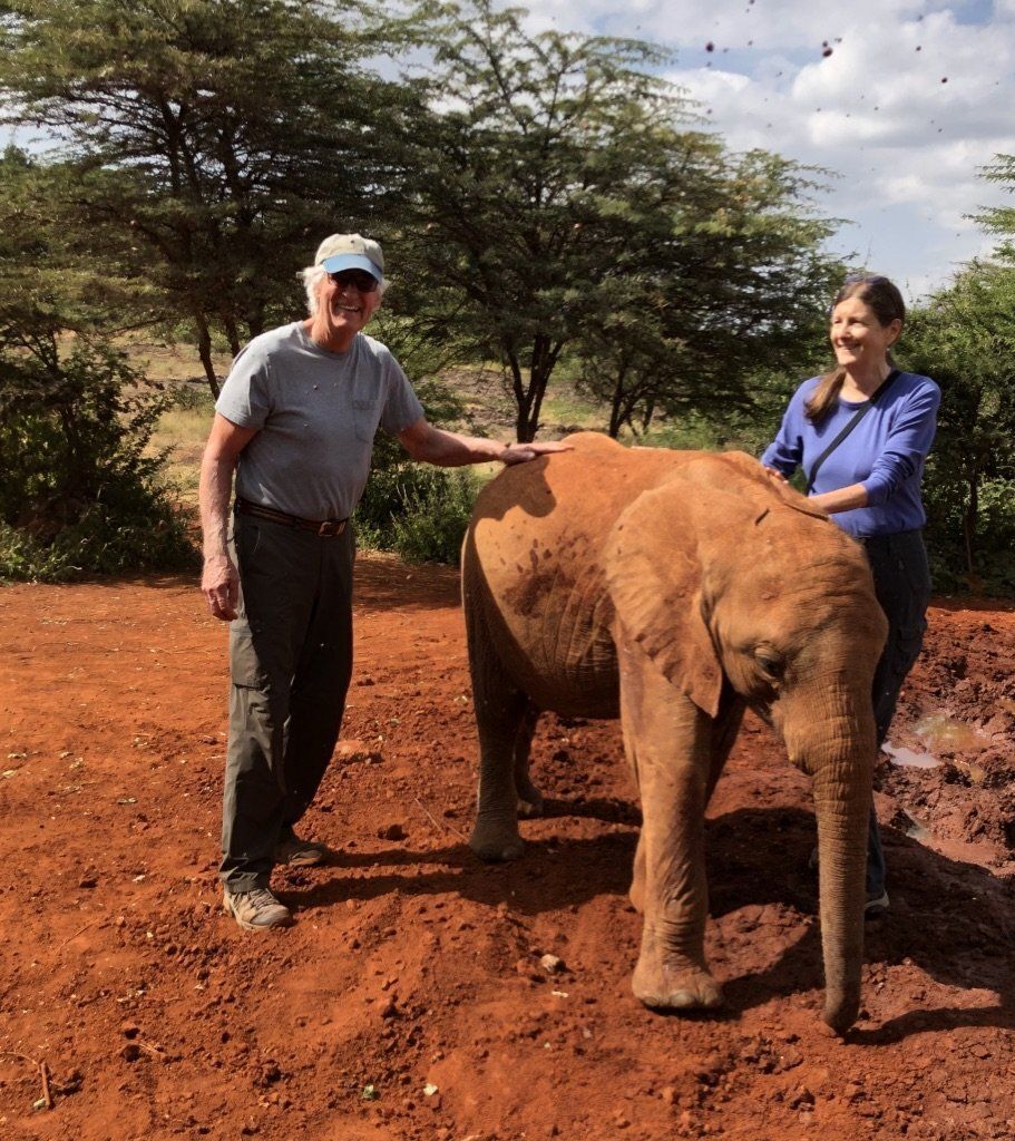 Two people standing with a small elephant on reddish soil with trees in the background.
