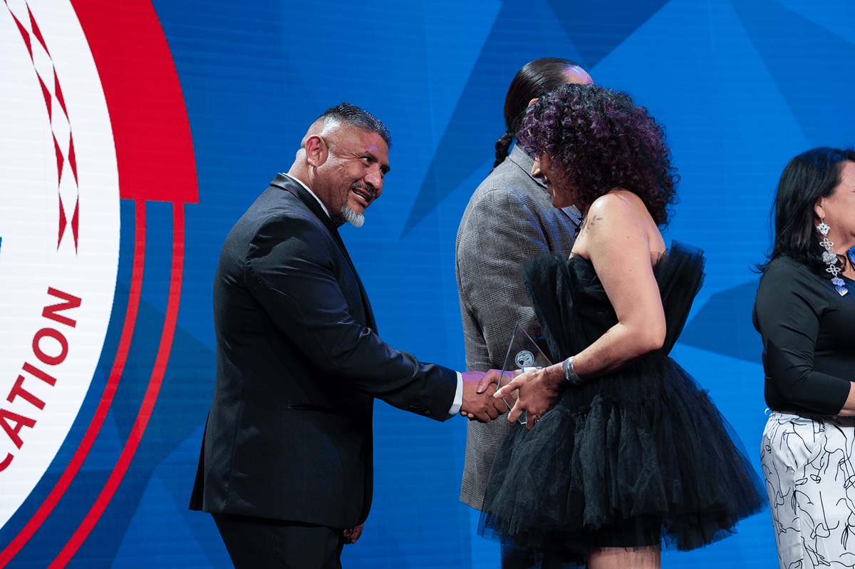 Two people shaking hands on stage, a man in a black suit and a woman in a black dress holding an award.