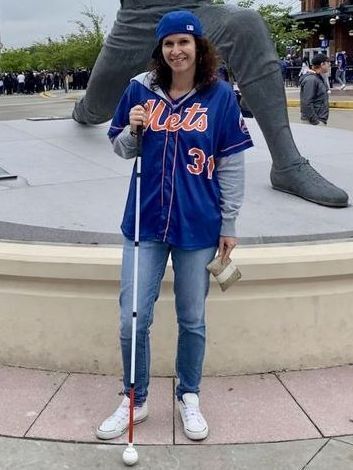 NY Ambassador Michele stands outside of Citi Field in NYC, home of the NY Mets baseball team. She is wearing a Mets shirt and baseball cap, holding her cane and standing in front of a statue of Tom Seaver, legendary pitcher of the NY Mets.