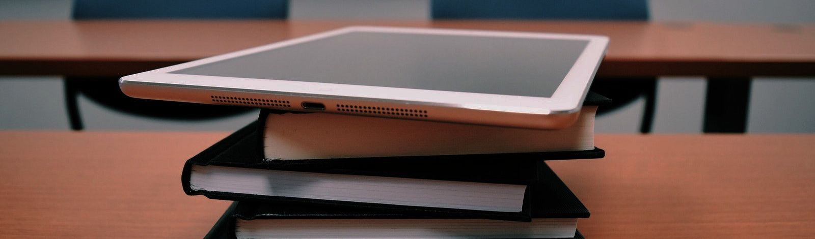 Sideview photo of a stack of books and a tablet set on a desk. The book covers are black and they are stacked offset from each other. A white tablet sits on top of the books, also offset. The desk is a red brown and in the background there is another desk