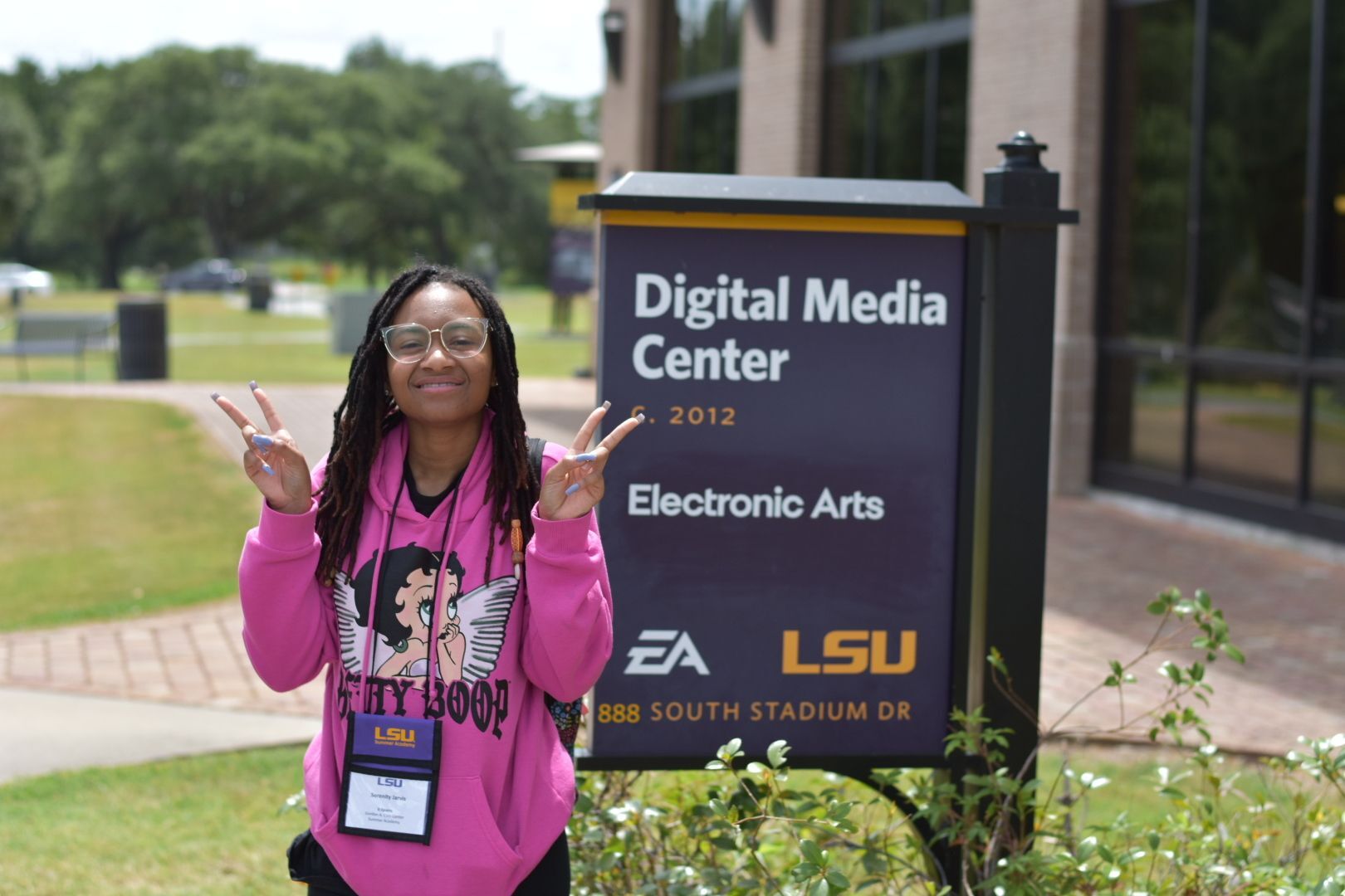 A student for the LSU Summer Academy posing happily with two peace signs in front of the Digital Media Center at LSU