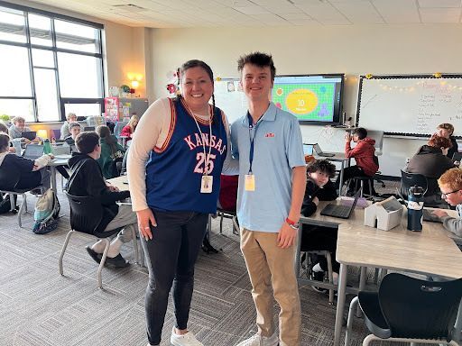 A smiling teacher wearing a Kansas basketball jersey stands next to a student intern in a classroom filled with middle school students working at tables. A colorful screen is visible on the smartboard behind them.