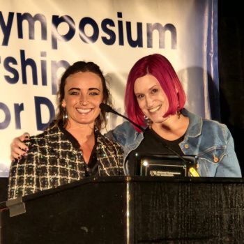 Two smiling women stand behind a podium in front of a conference banner. The left has dark hair and a patterned blazer; the right has bright pink hair and a denim jacket, holding a black-framed award plaque.