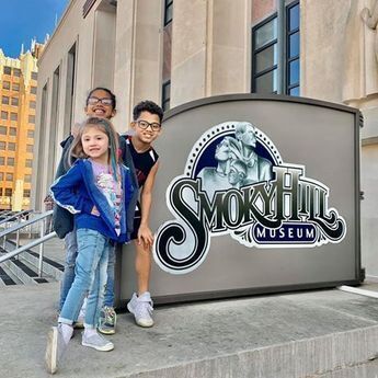 Three Children posing with the exterior sign for the Smoky Hill Museum.
