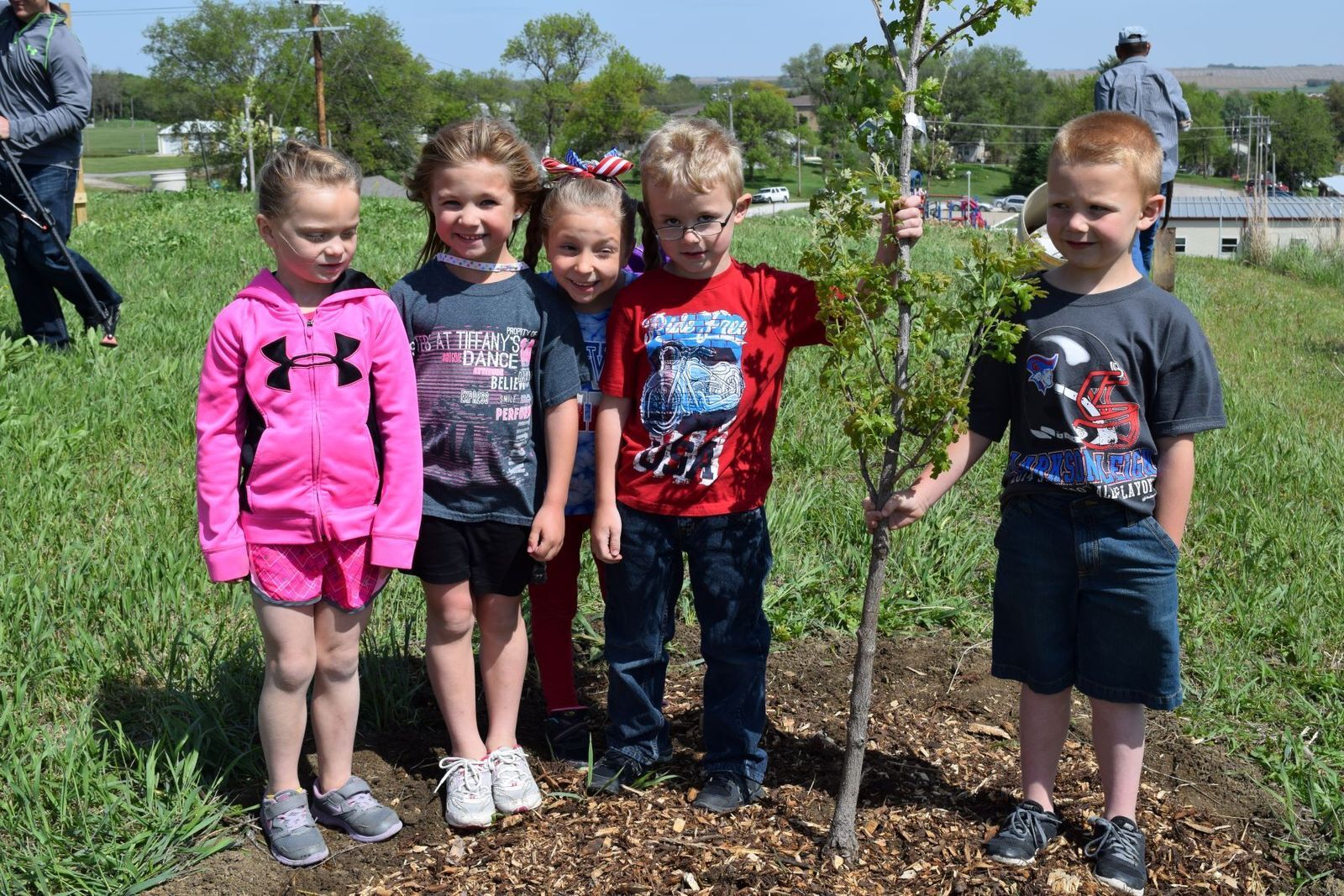 A group of five young children pose with a newly planted small tree.