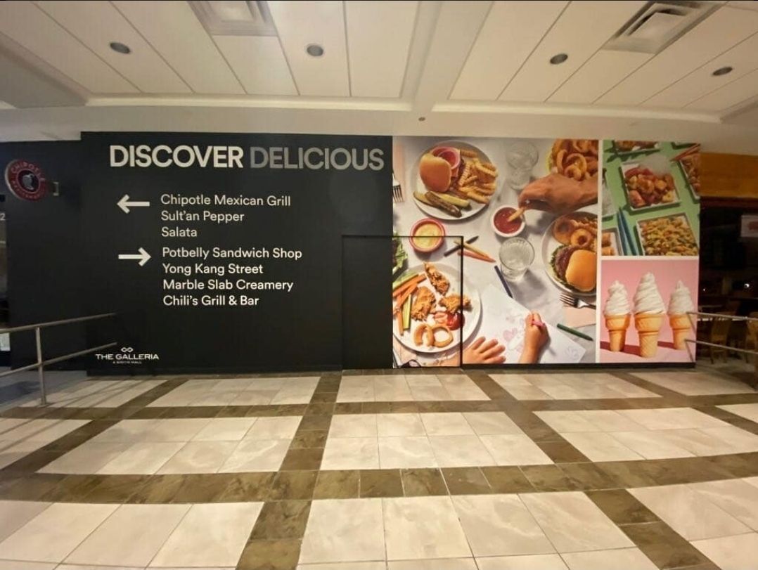 Large barricade wrap inside The Galleria mall displaying “Discover Delicious” with directional signage and vibrant food photography, promoting nearby restaurants like Chipotle, Potbelly, and Marble Slab.