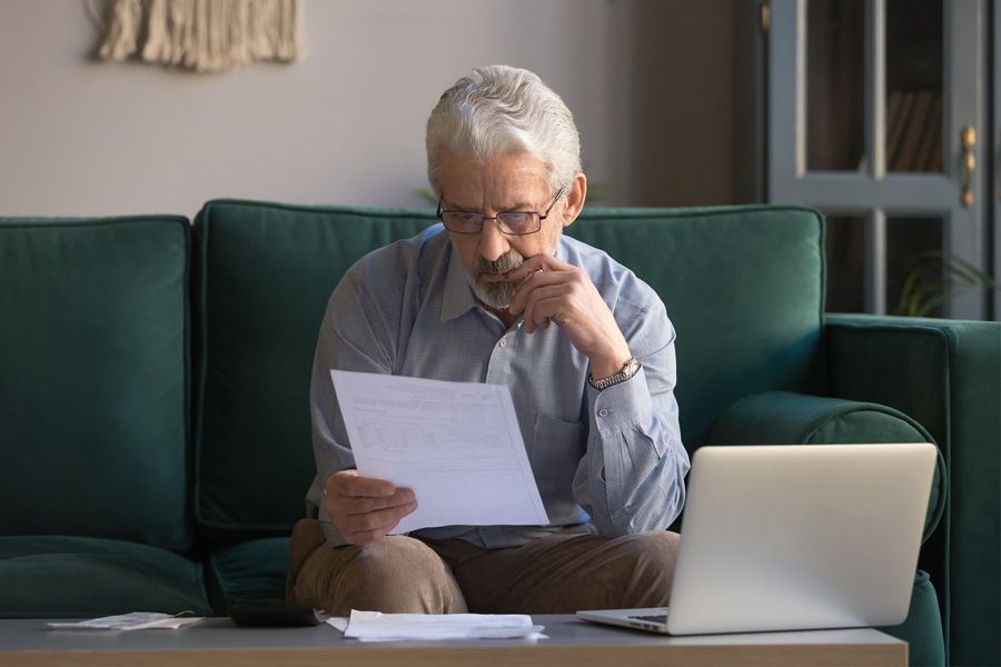 Older man sitting on his couch in front of his laptop, holding and focusing on an important paper