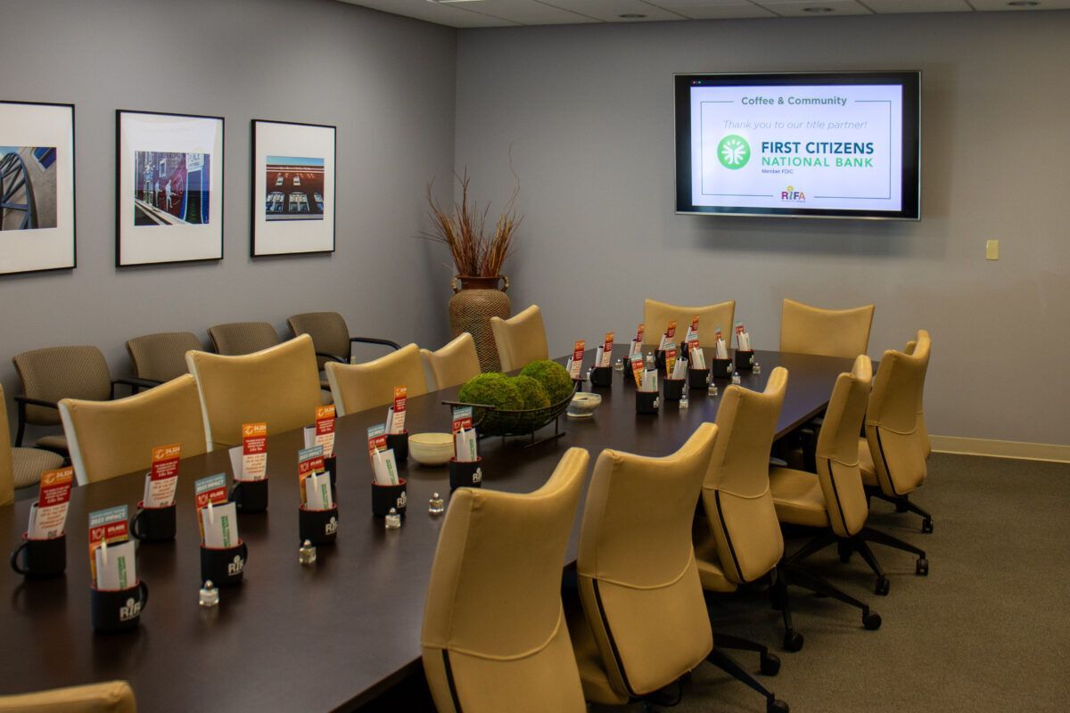 A conference room with a large table, beige chairs, a flat-screen display, and framed photos on the wall.