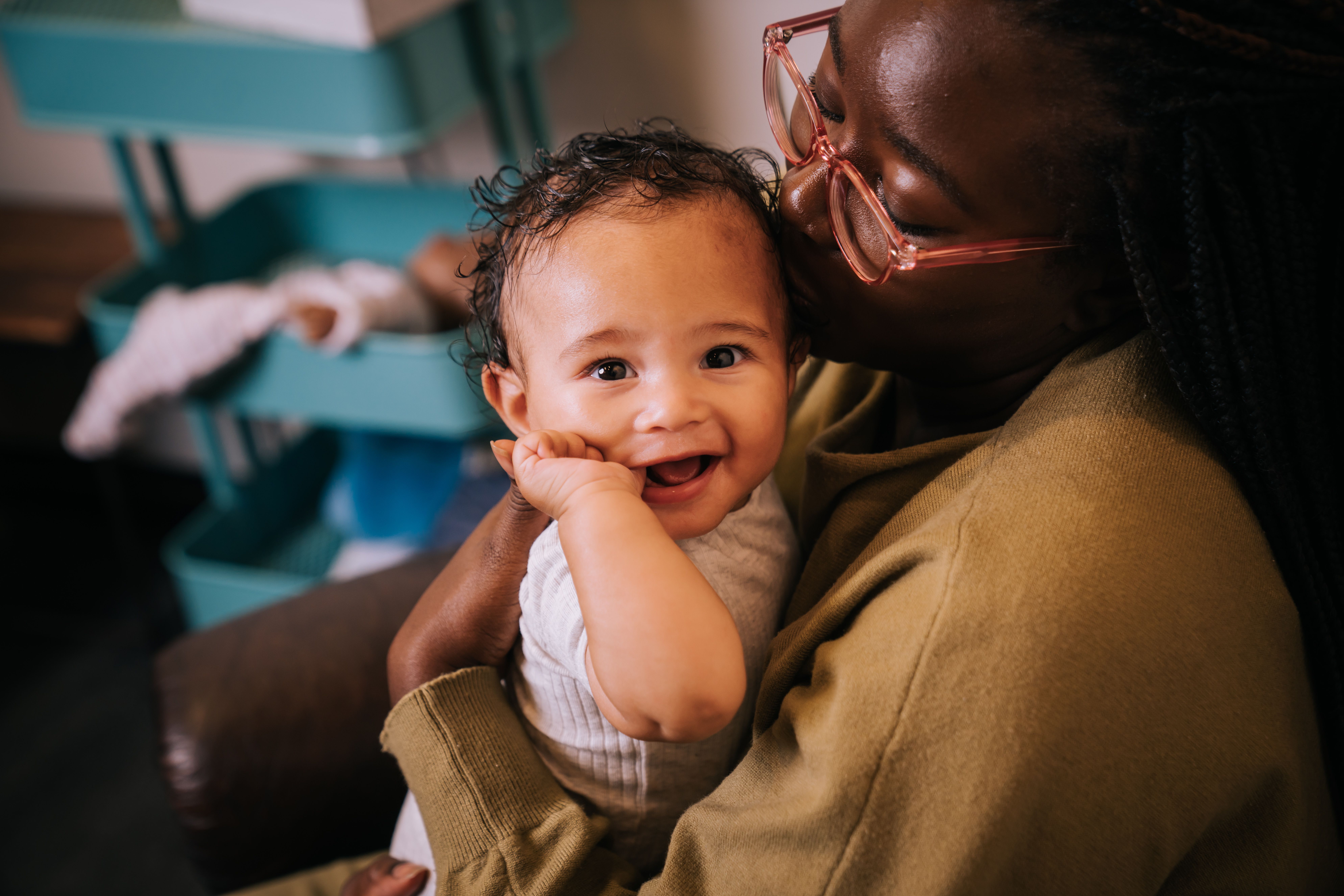 Black mother kissing her black baby on the temple. Baby is smiling at the camera with one thumb in his mouth