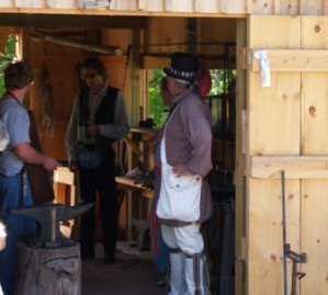 Three people in a wooden workshop, with an anvil and historical attire.