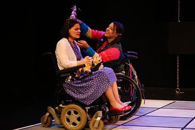 POWER PLAYS 2014. (L to R): Ann Marie is wearing a purple dress and white sweater. Jamie is wearing a multicolor outfit. They are in wheelchairs and Jamie is placing a party hat on Ann's head.