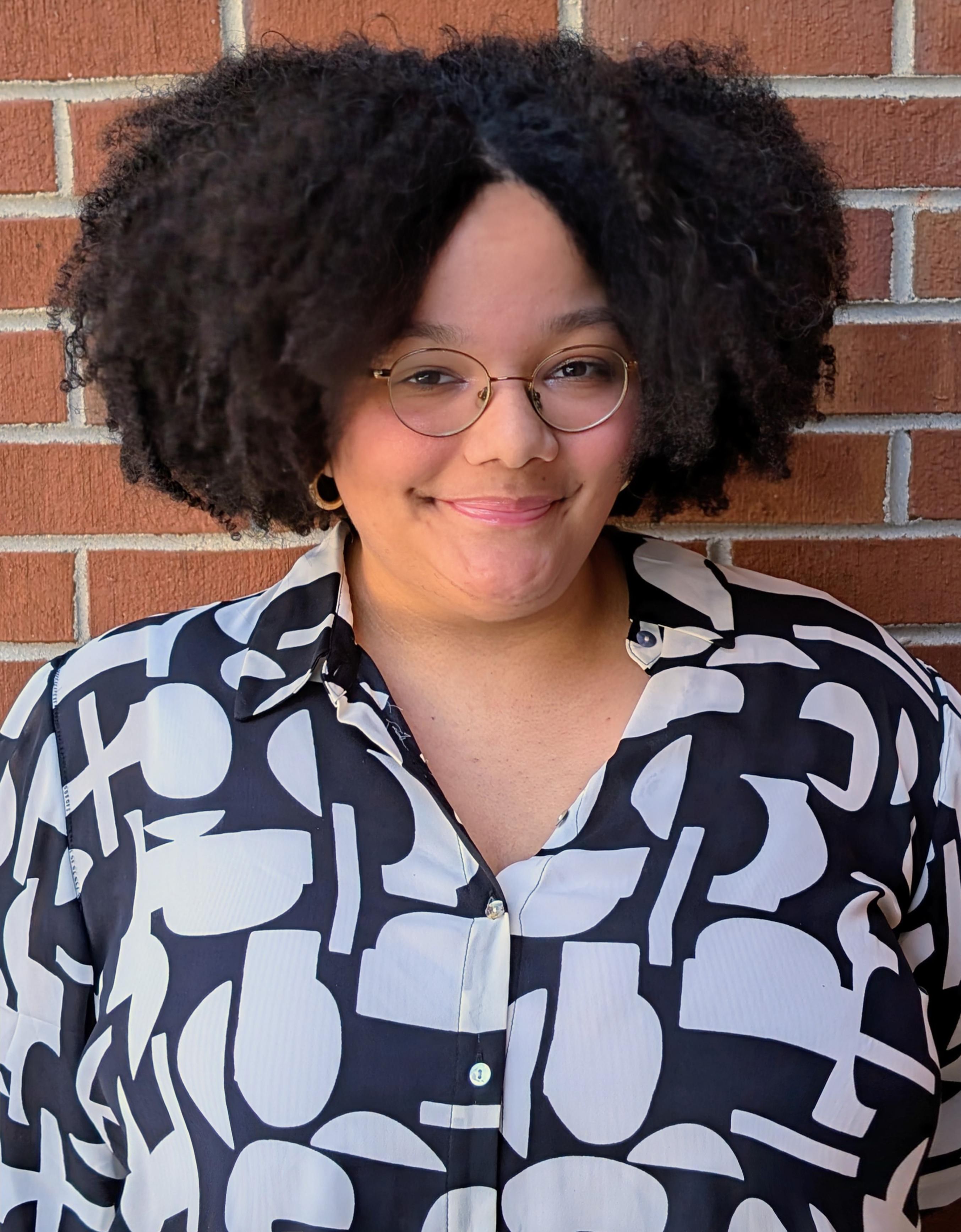 "A smiling person with round glasses and curly hair stands in front of a brick wall. They are wearing a black and white patterned button-up shirt.