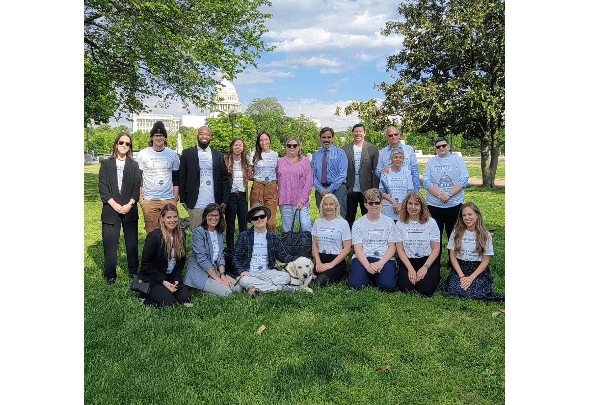 A group photo with people outside on the grass. The US Capitol building is the background. There's one person with a guide dog and a few others with their white canes. Everyone wears a matching white t-shirt that says "Ask me about USH"