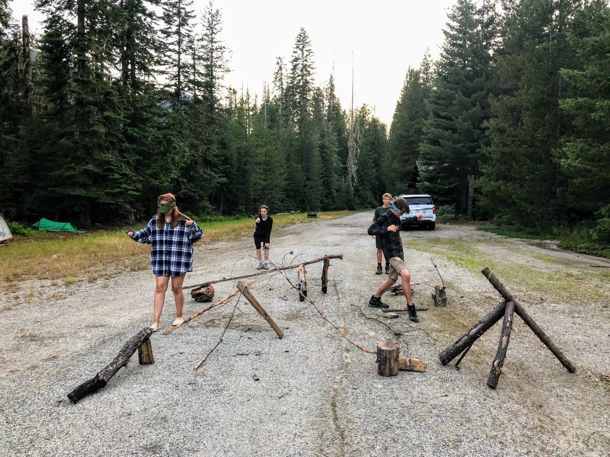 [Image Description: A group of four MCC Youth Members are visibly blindfolded, walking around cautiously in an obstacle course made of found items in the forest; i.e., sticks, logs, rocks, and more. Behind them, there is a tent and an MCC vehicle parked.]