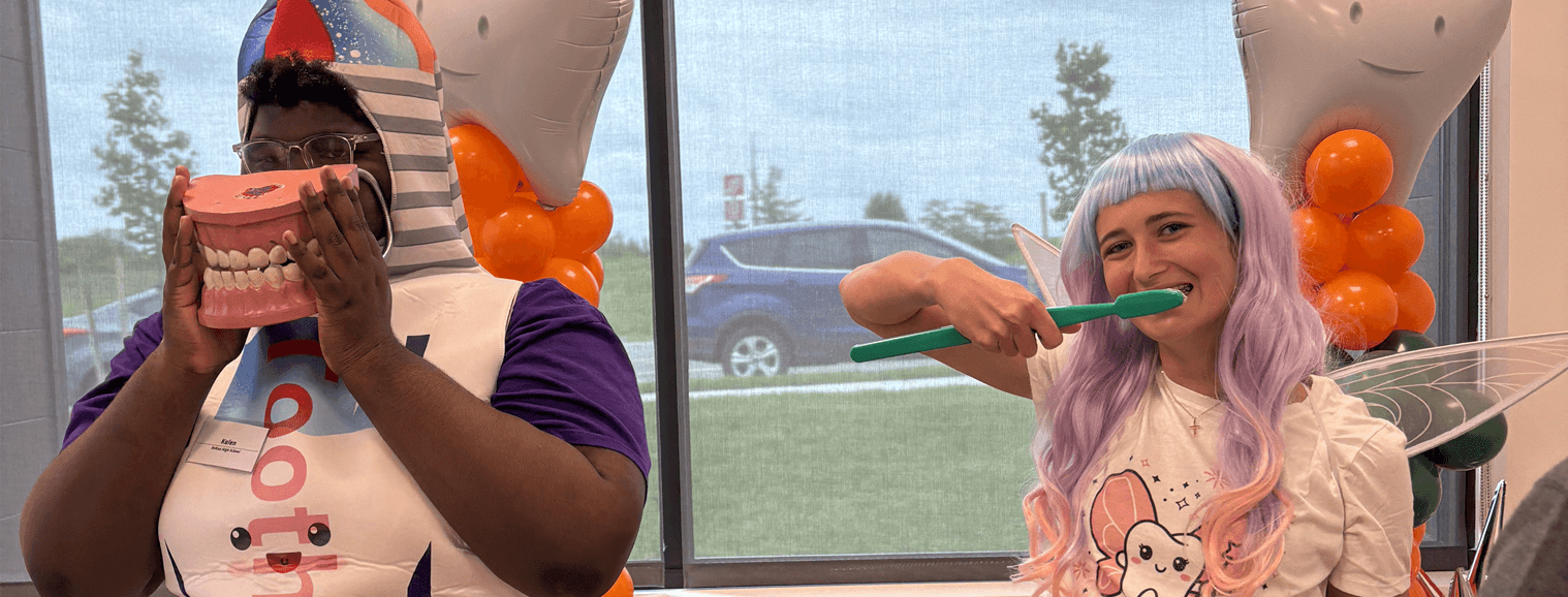 young man dressed up as toothpaste holding tooth model and young woman dressed as tooth fairy brushing teeth