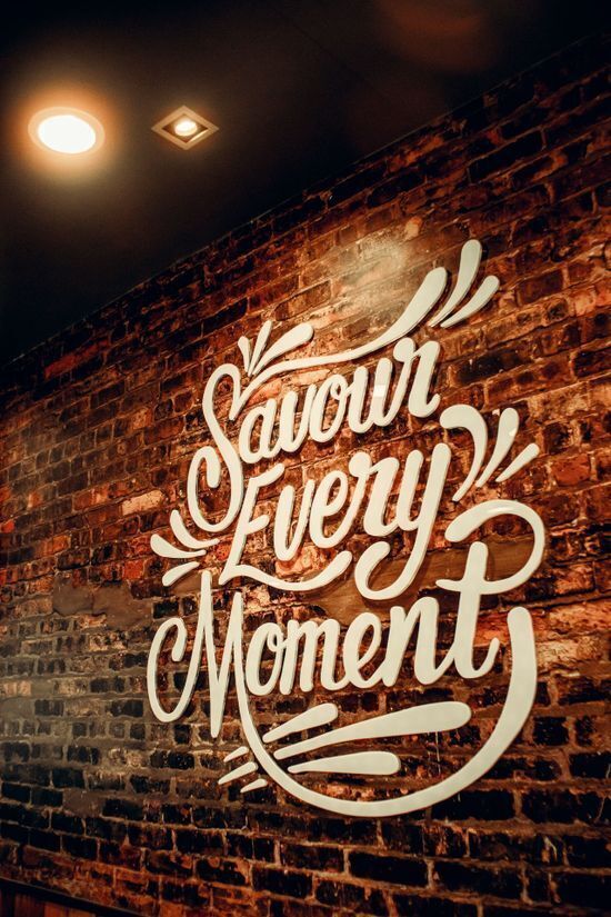 Retail wall graphics reading "Savour Every Moment" in white script lettering mounted on an exposed brick wall with warm lighting.