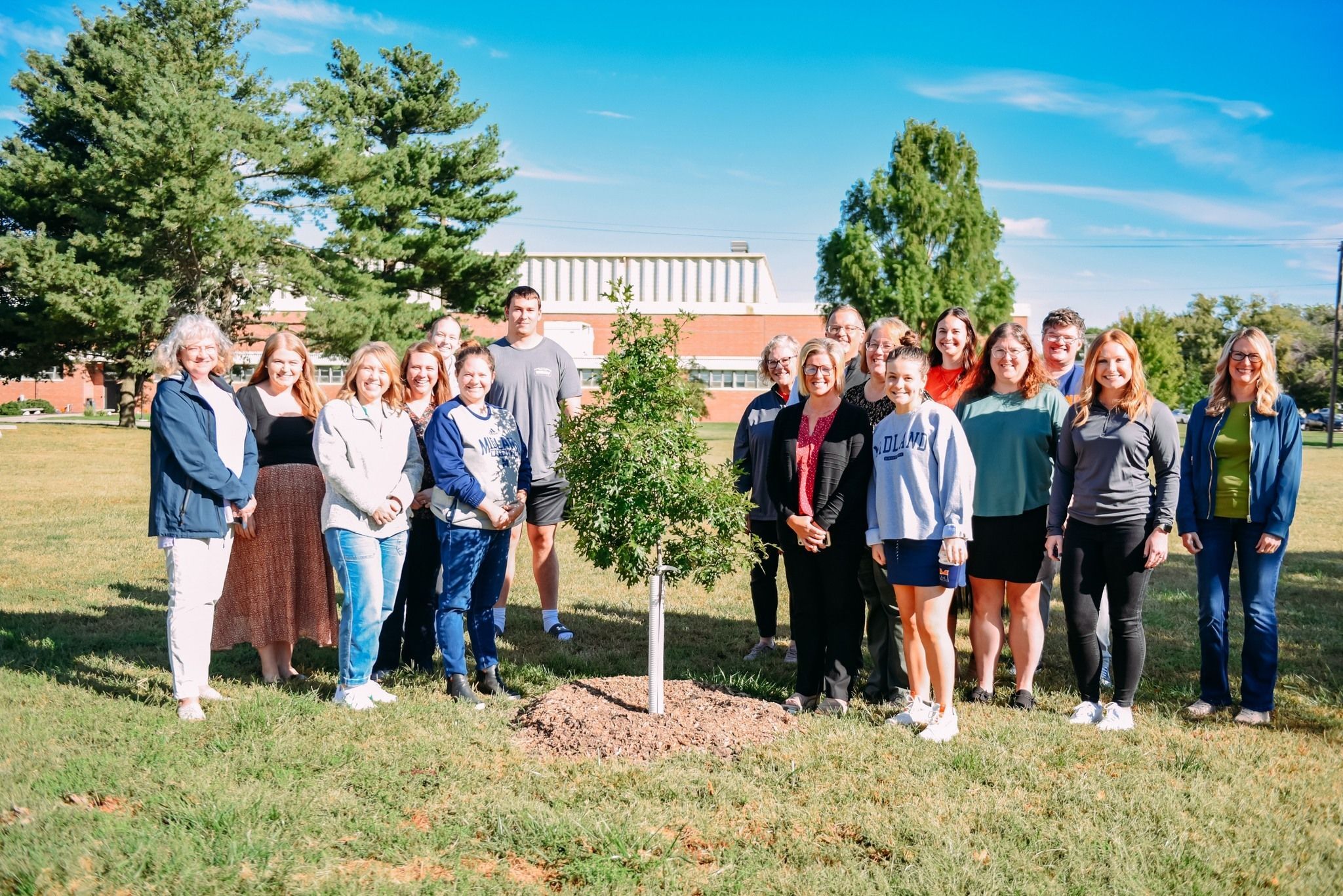 A group of people stand next to a newly planted tree. 