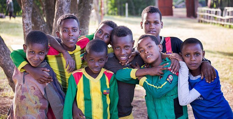 A group of eight smiling children standing closely together outdoors under trees.