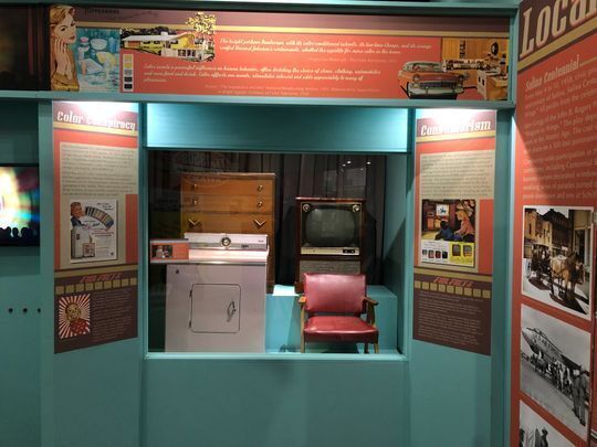 Exhibit case at the Smoky Hill Museum showing two a 1950s pink clothes dryer, console TV, blonde wood chair with red vinyl seat and back and bedroom highboy dresser.