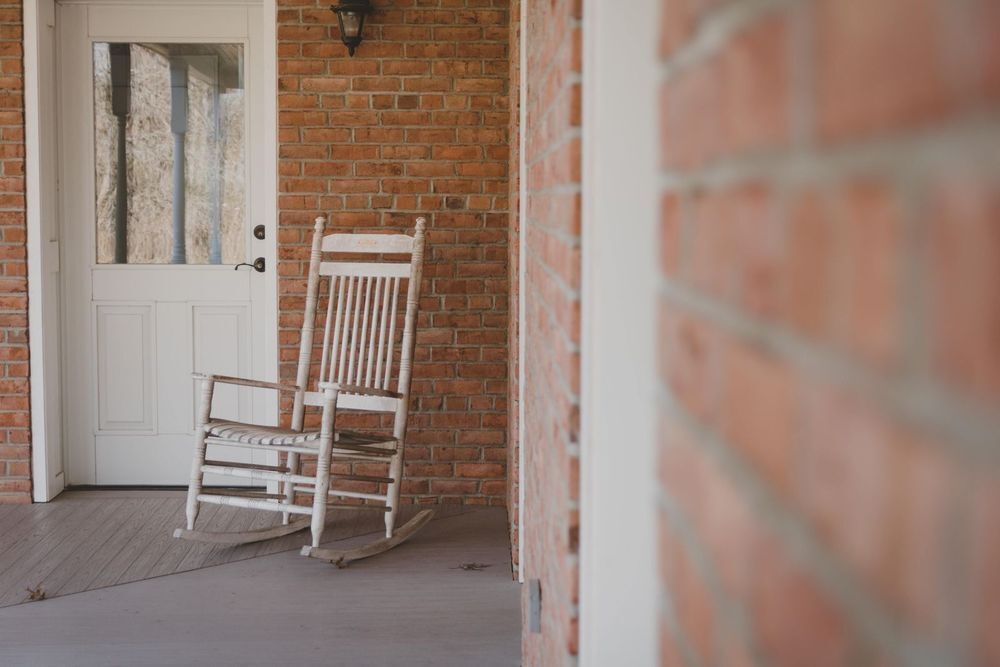 A close up shot of the exterior of a brick house with worn floor boards on the front porch, with a white rocking chair in focus. A door is also visible with the reflection of some trees and the porch balusters in the glass. 