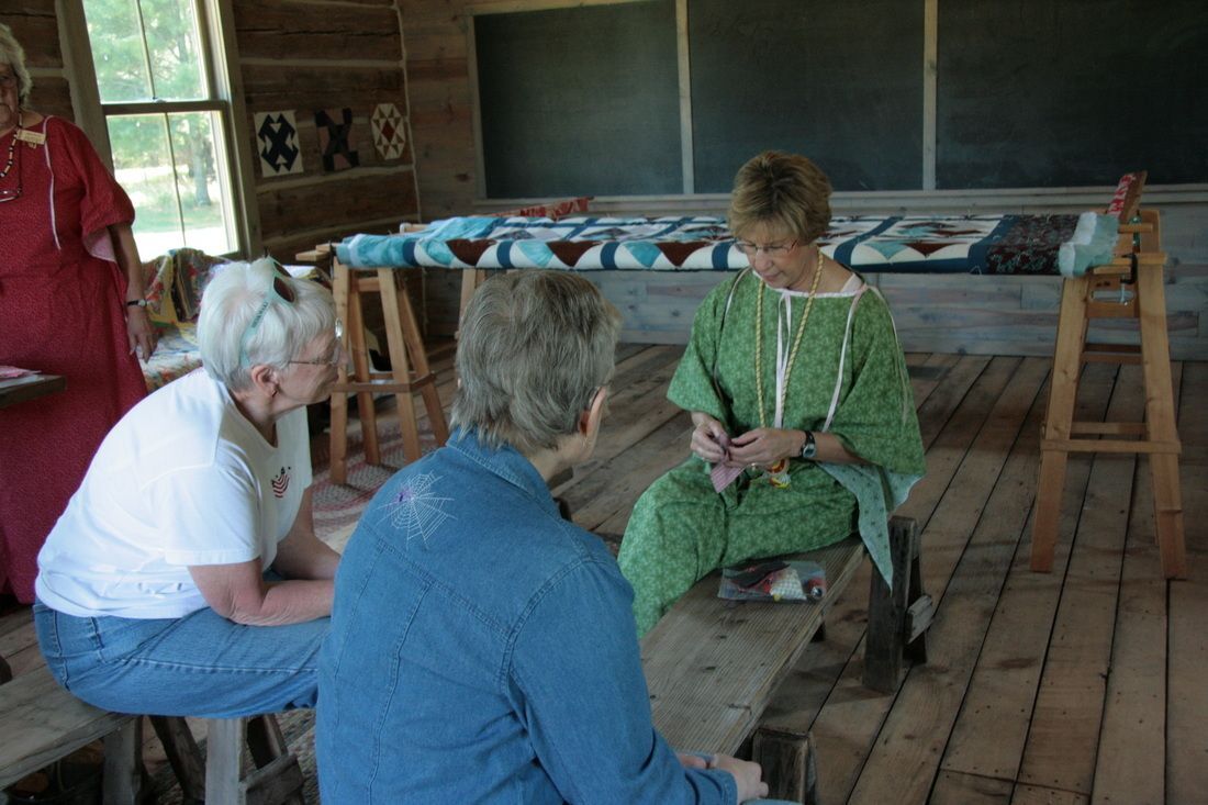 A group of people in a rustic room with a woman in a green dress engaged in needlework.