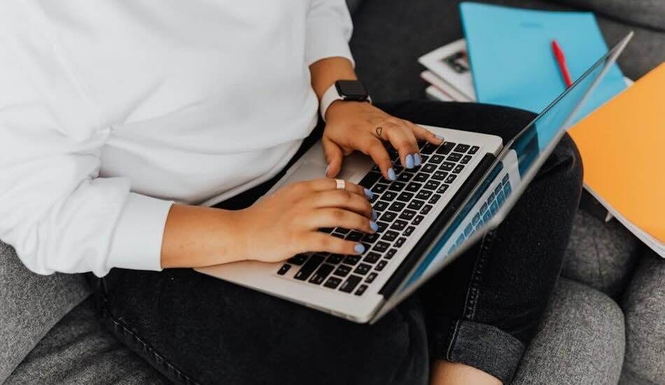 Photo showing a person typing on a laptop, seated on a grey couch and wearing dark jeans and a white shirt.