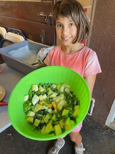A 9 or 10 year old girl looks up while holding her bright green bowl of freshly cooked squash, green beans, and green peppers for the photographer to admire