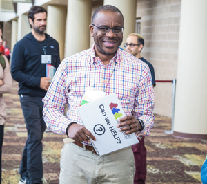 A man smiling at the camera and holding a sign that says Can I Help? Behind him are a few other conference goers.