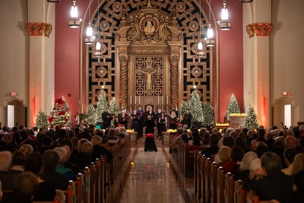 Seraphic Fire singers perform for a full audience on a church alter in front of Christmas trees strung with white lights.