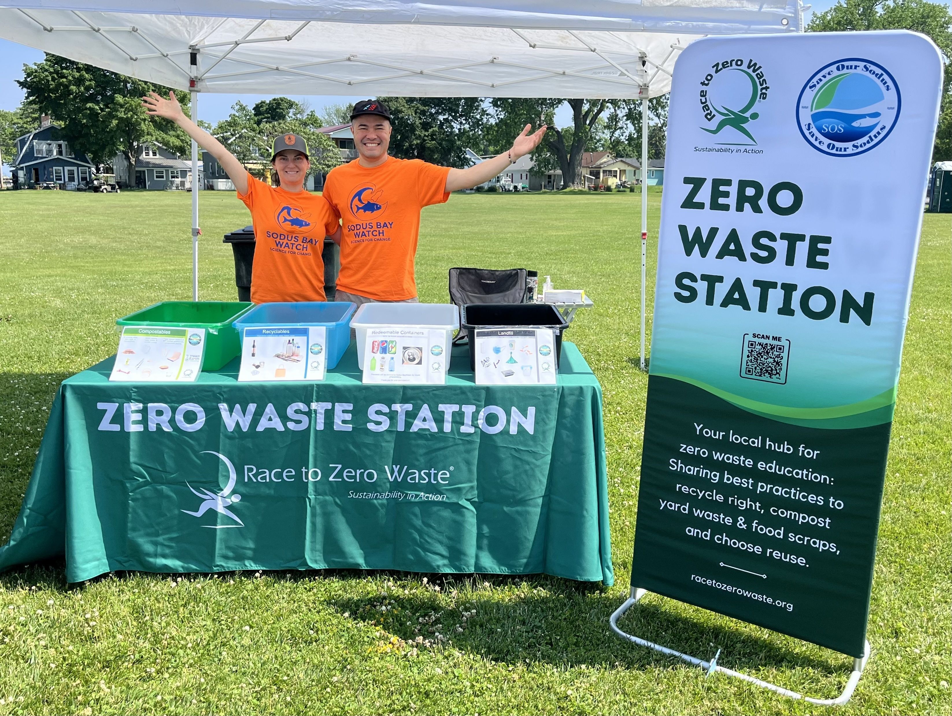 The Zero Waste Station Pop-Up used by volunteers with Save Our Sodus. Two volunteers raise their arms behind the Zero Waste Station table, with the standing banner to the right.