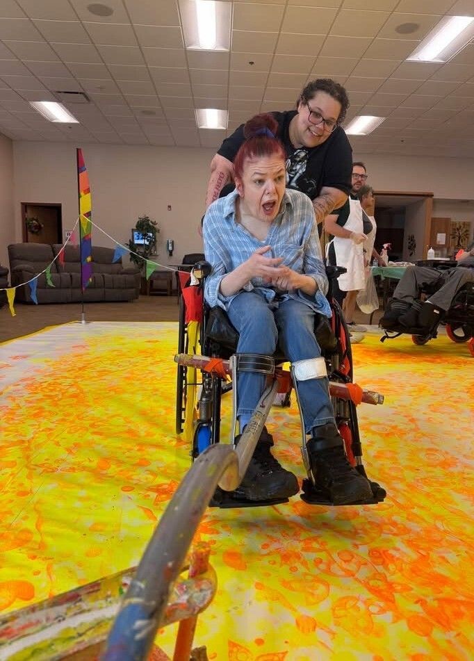 A woman in a wheelchair has an excited expression.  She is pushed by a woman behind her, smiling.  They are on a large, colorfully painted canvas and there is an adaptive paint roller attached to the front of the wheelchair.