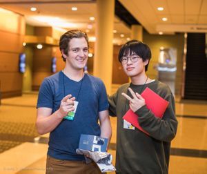 Two young men stand looking at the camera and smiling. One is pointing, and one is holding up a peace sign.