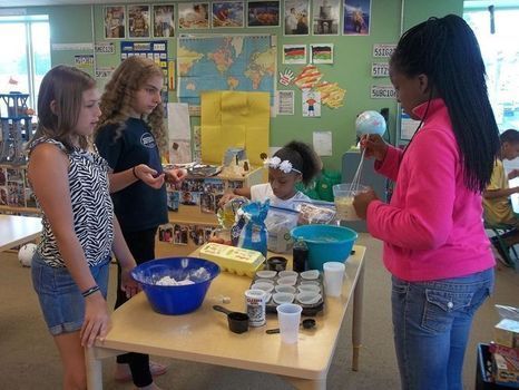A group of students stand around a table while mixing ingredients together in a bowl, such as eggs and flour.