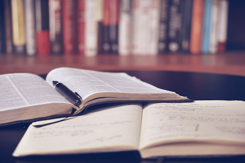 Open reference book and open journal on dark wooden table with a fountain pen.  Book shelf in the background.