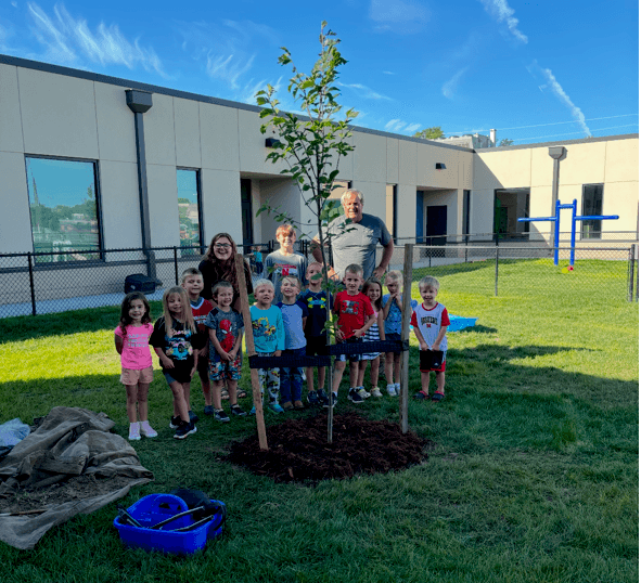 A group of elementary students stand around a newly planted tree in their schoolyard