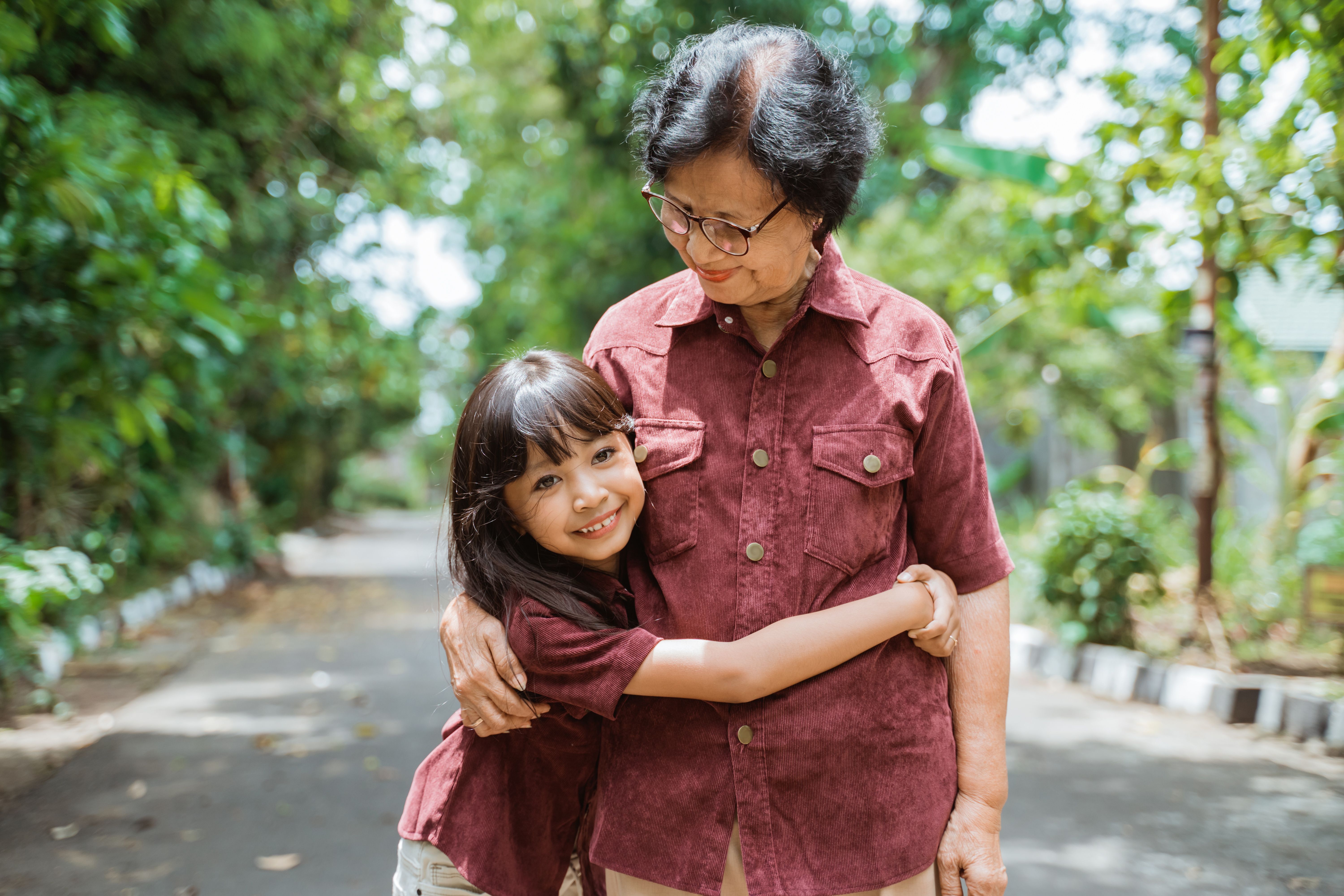 Photo of grandma and granddaughter of Asian descent hugging on a sunny day against a backdrop of trees.  