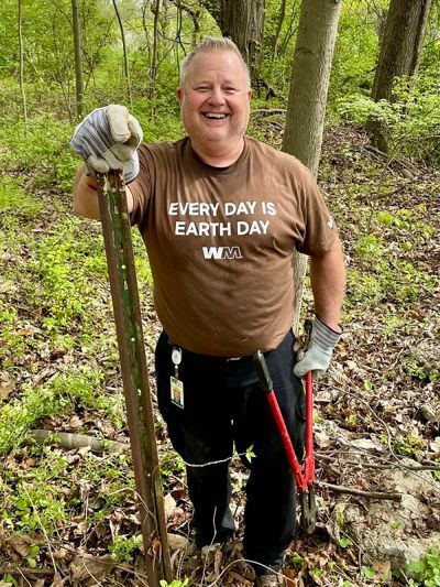 Large man smiles widely at the camera as he hangs on to a metal fencepoint.  He's wearing a brown T-Shirt that says "EVERY DAY IS EARTH DAY" and he is holding a pair of bolt cutters in his left hand.