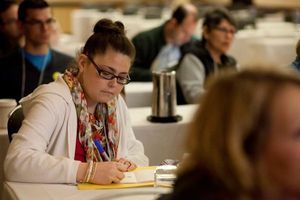 A woman with a bun and glasses is sitting at a conference table taking notes. You can see other conference goers behind her.