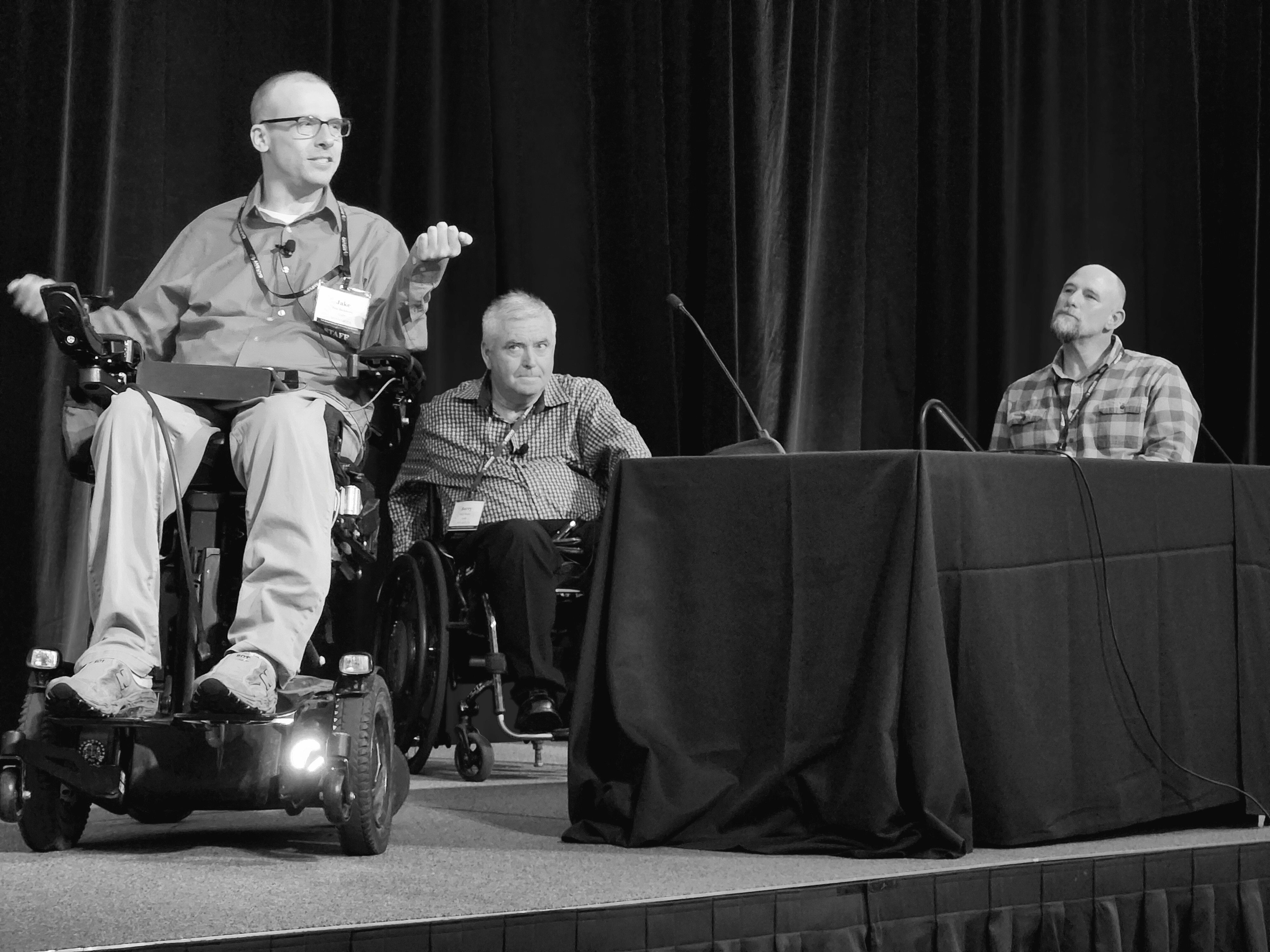 White man in wheelchair on a stage with two other white men in wheelchairs behind him
