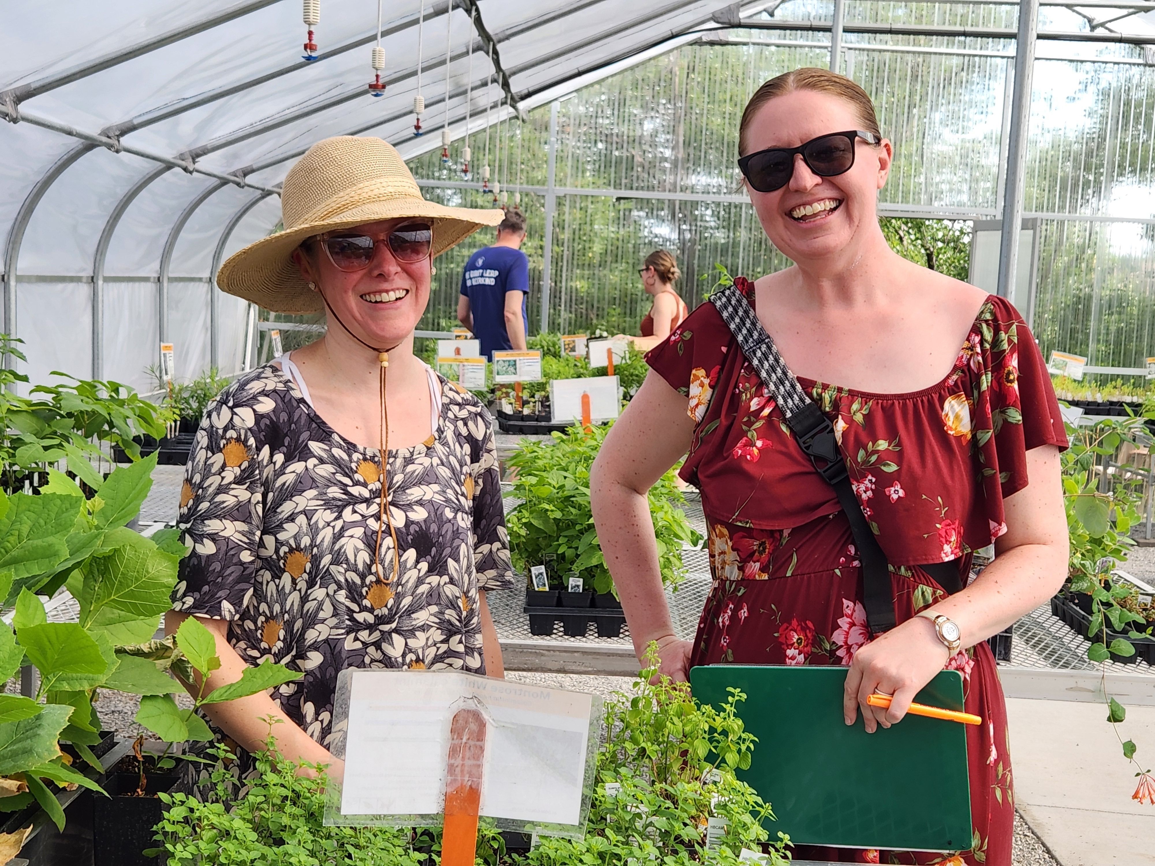 A woman wearing a floral shirt and a wide-brimmed hat and a woman wearing a red floral dress stand side-by-side in a greenhouse. 