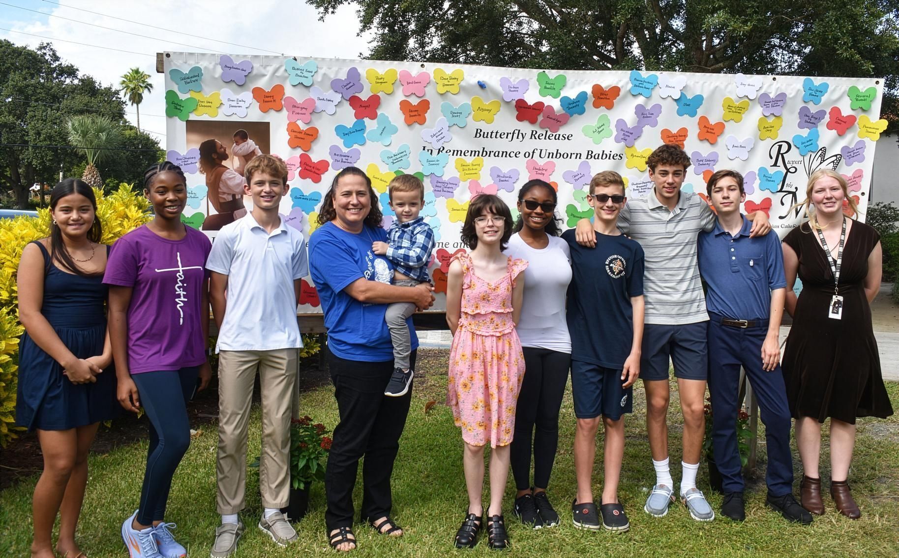 Standing in front of the butterfly release memorial is St. Helen School Principal Debbie Irish, holding grandson Dominic, with student volunteers at the Vero Beach parish Oct. 26, 2025.