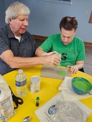 2 men sit at a table where 1 helps the other to squeeze green paint out of a bottle