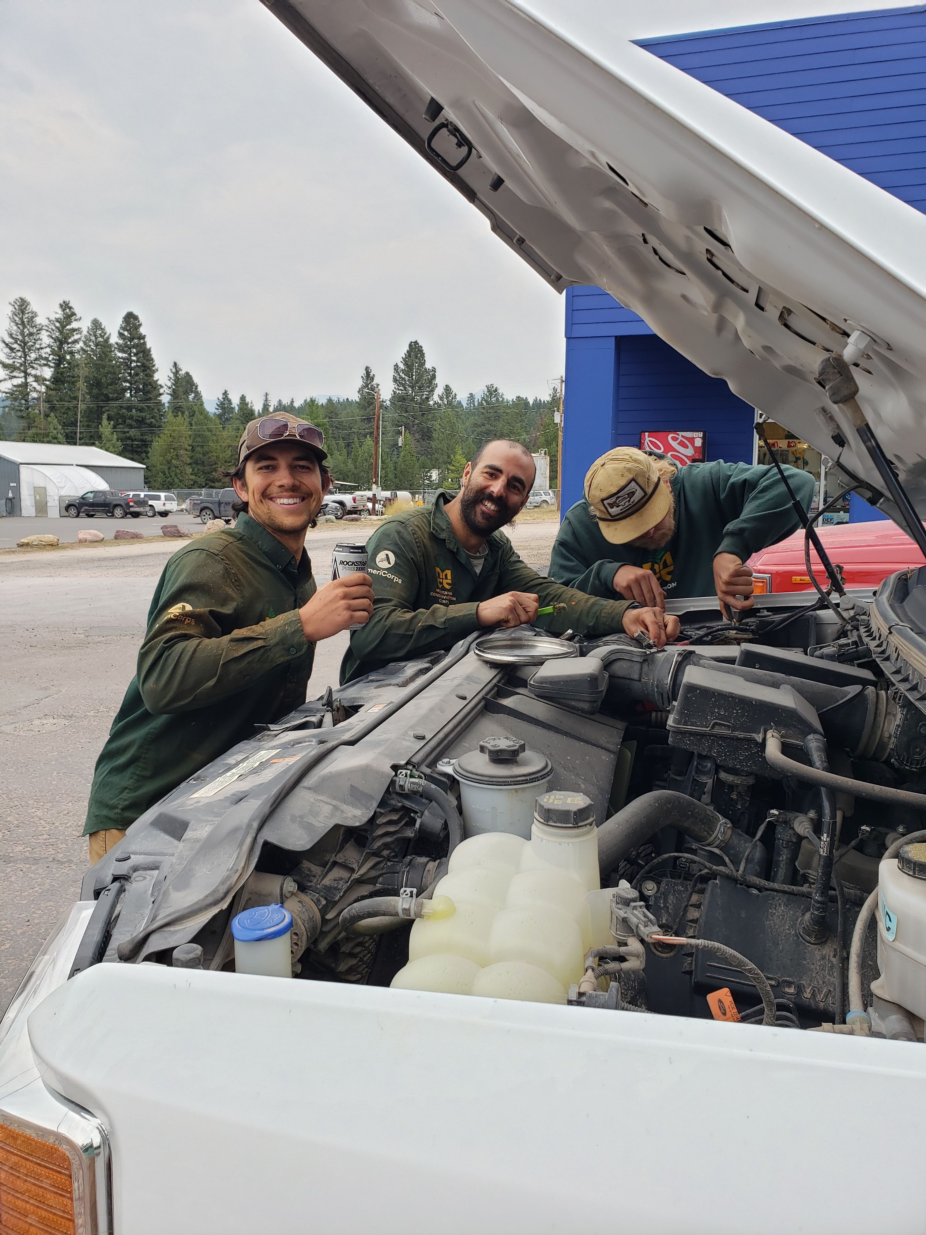 Three crew members huddle around the front end of a truck, hood popped as they assess the problem.