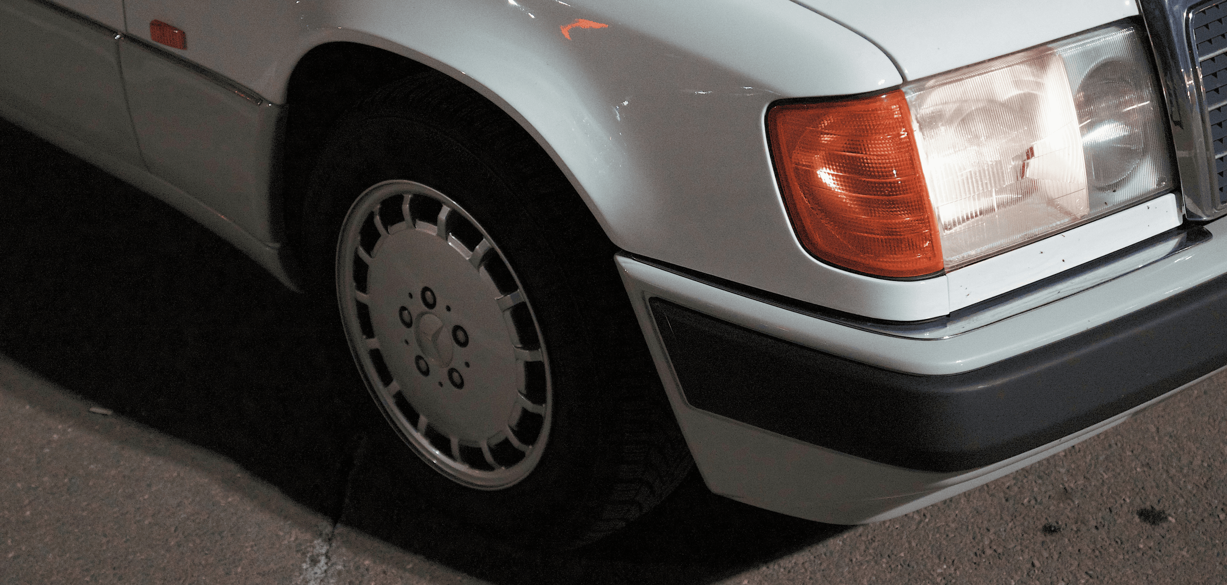 A close-up photo of a dated white vehicle, showing some of it's front bumper, headlight, and front wheel