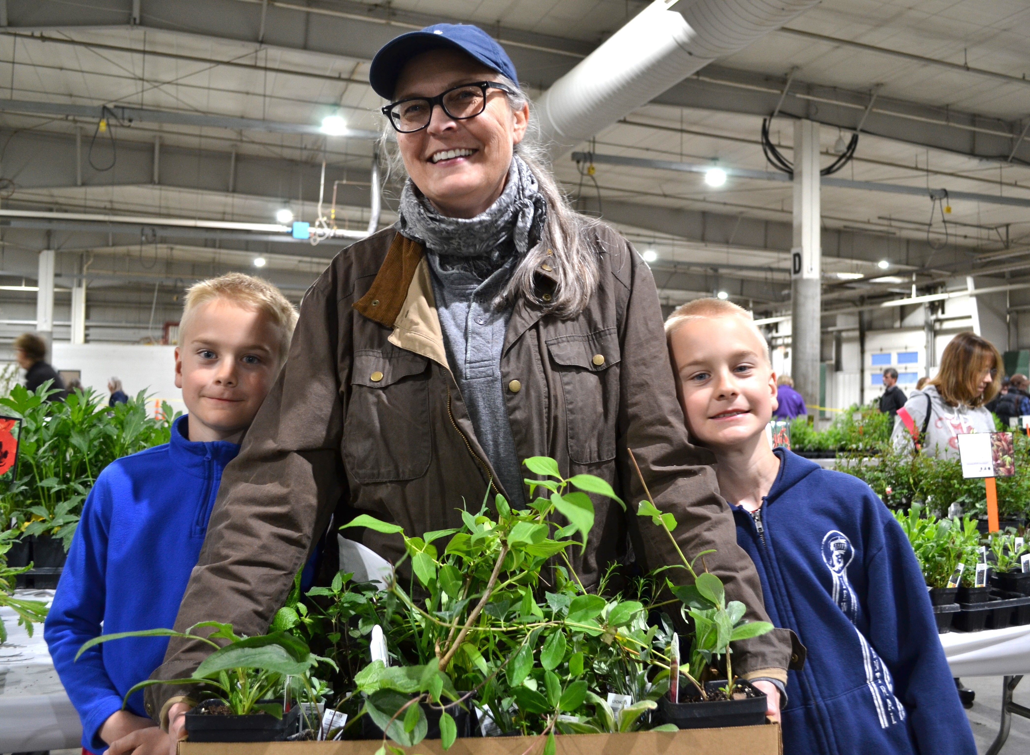 A mom wearing a blue baseball cap holds a cardboard flat of plants with her twin boys wearing blue sweatshirts on either side of her. 